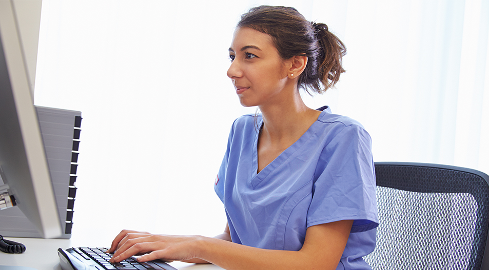 Female nurse sat at desk typing into keyboard and looking at desktop screen
